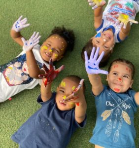 Niños acostados sobre grama, sonrientes y mostrando las manos pintadas de colores.